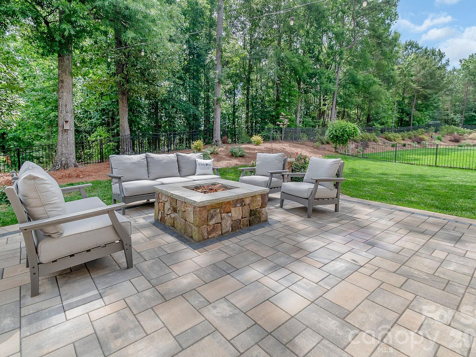 The custom paver patio with stone-rimmed fire pit, four chairs gathered around it, lawn rolling toward the tree line.