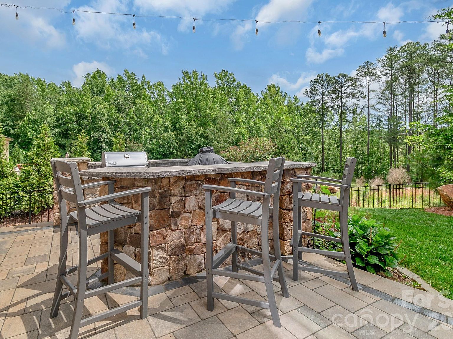 The outdoor kitchen: stacked stone counter, built-in grill, bar seating for three under string lights.